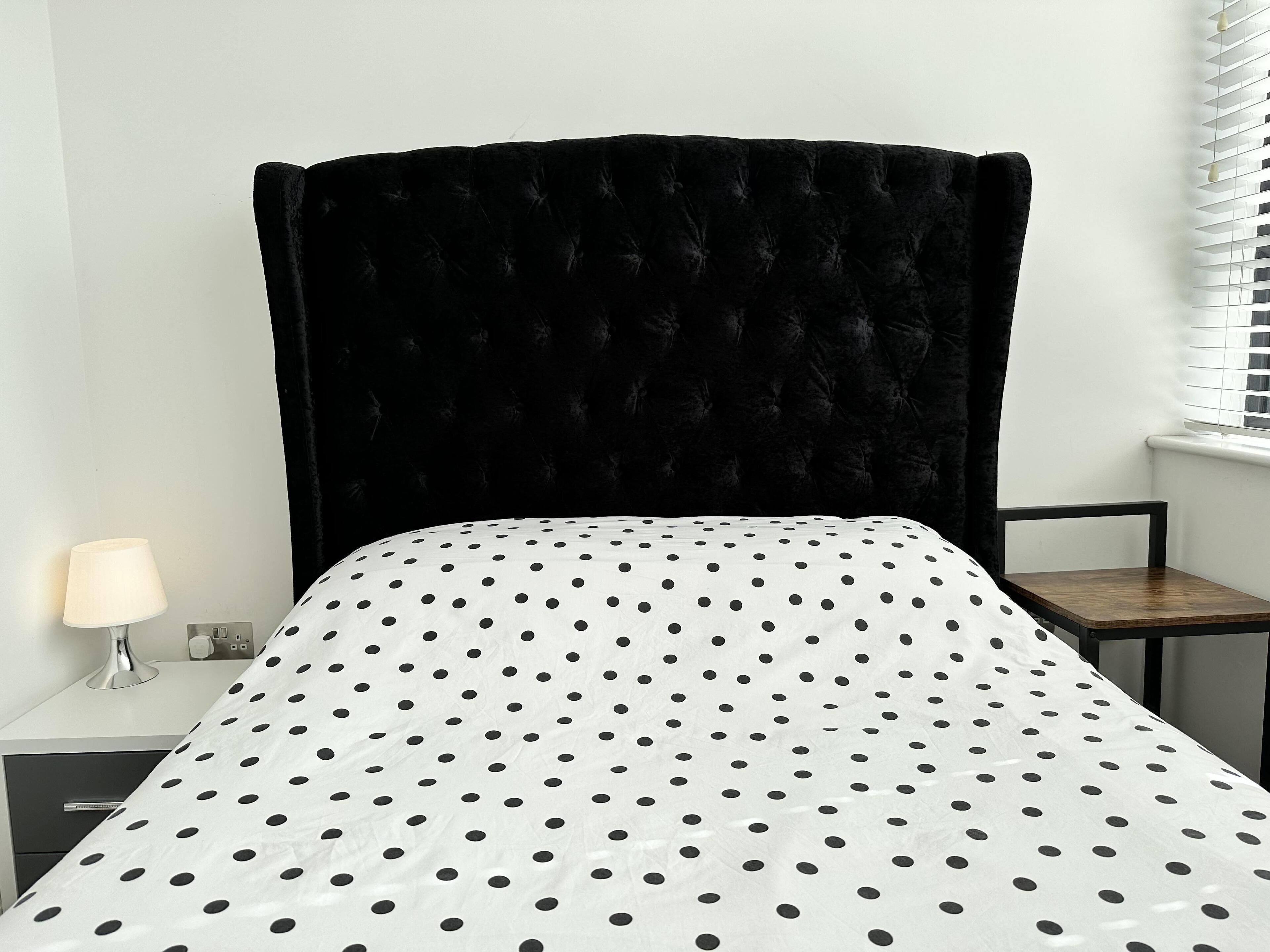 Un dormitorio con cabecera tapizada en negro, colcha blanca con lunares negros, mesita de noche con lámpara y una silla.