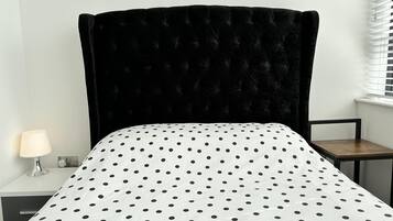 Un dormitorio con cabecera tapizada en negro, colcha blanca con lunares negros, mesita de noche con lámpara y una silla.