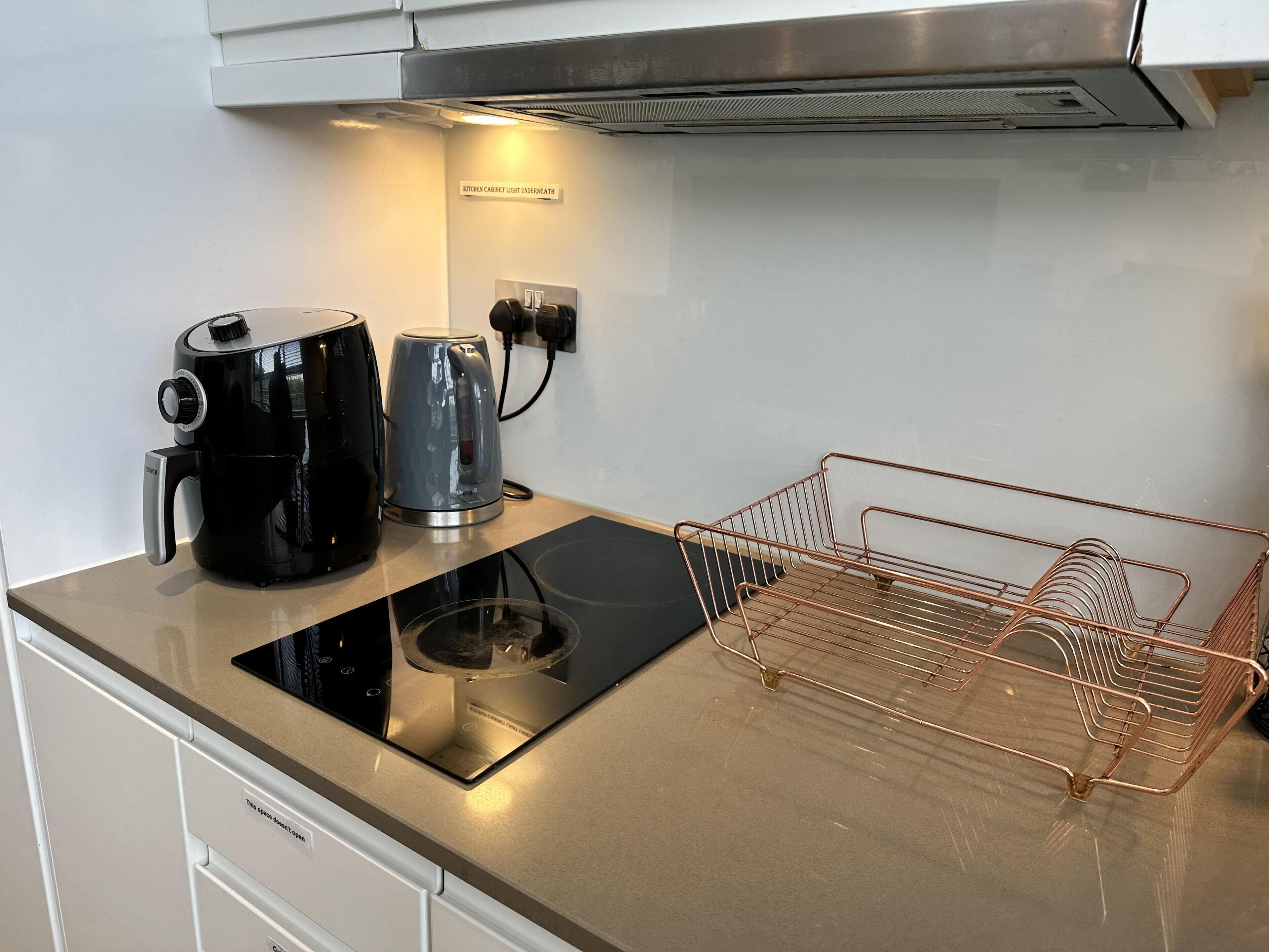 Una cocina con una cafetera negra, un hervidor plateado y una cesta de alambre de cobre sobre una encimera de acero inoxidable.