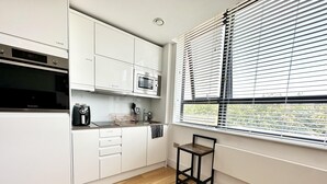 A modern kitchen with white cabinetry, a built-in oven, and a window with blinds. - Condo Apartment With Workspace (Haywards Heath)