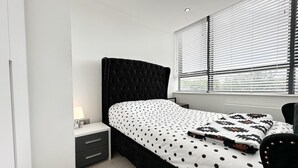 A bedroom with a black upholstered headboard, a bed with a white and black polka dot bedspread, a bedside table with a lamp, and a window with blinds. - Condo Apartment With Workspace (Haywards Heath)