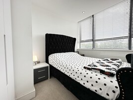 A bedroom with a black upholstered headboard, a bed with a white and black polka dot bedspread, a bedside table with a lamp, and a window with blinds.