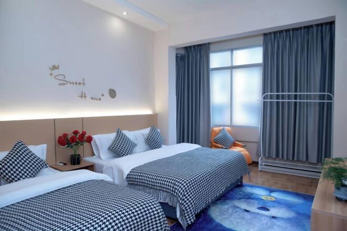 A hotel room with two beds, a nightstand with a vase of red roses, a window with curtains, and a wall with a decorative sign.