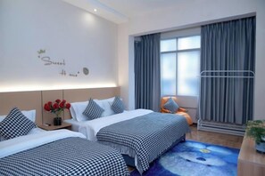 A hotel room with two beds, a large window with curtains, a bedside table with a vase of red flowers, and a wall with a decorative phrase.