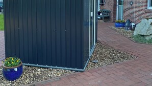 Eine Metallschuppen mit geneigtem Dach, ein Backsteinhaus mit Fenster und Tür, und ein gepflasterter Weg.