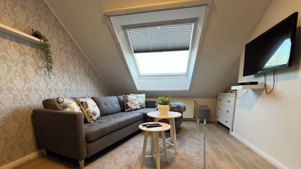 Ein modernes Wohnzimmer mit einem grauen Sofa, einem kleinen runden Tisch, einer Topfpflanze und einem an der Wand befestigten Fernseher.
