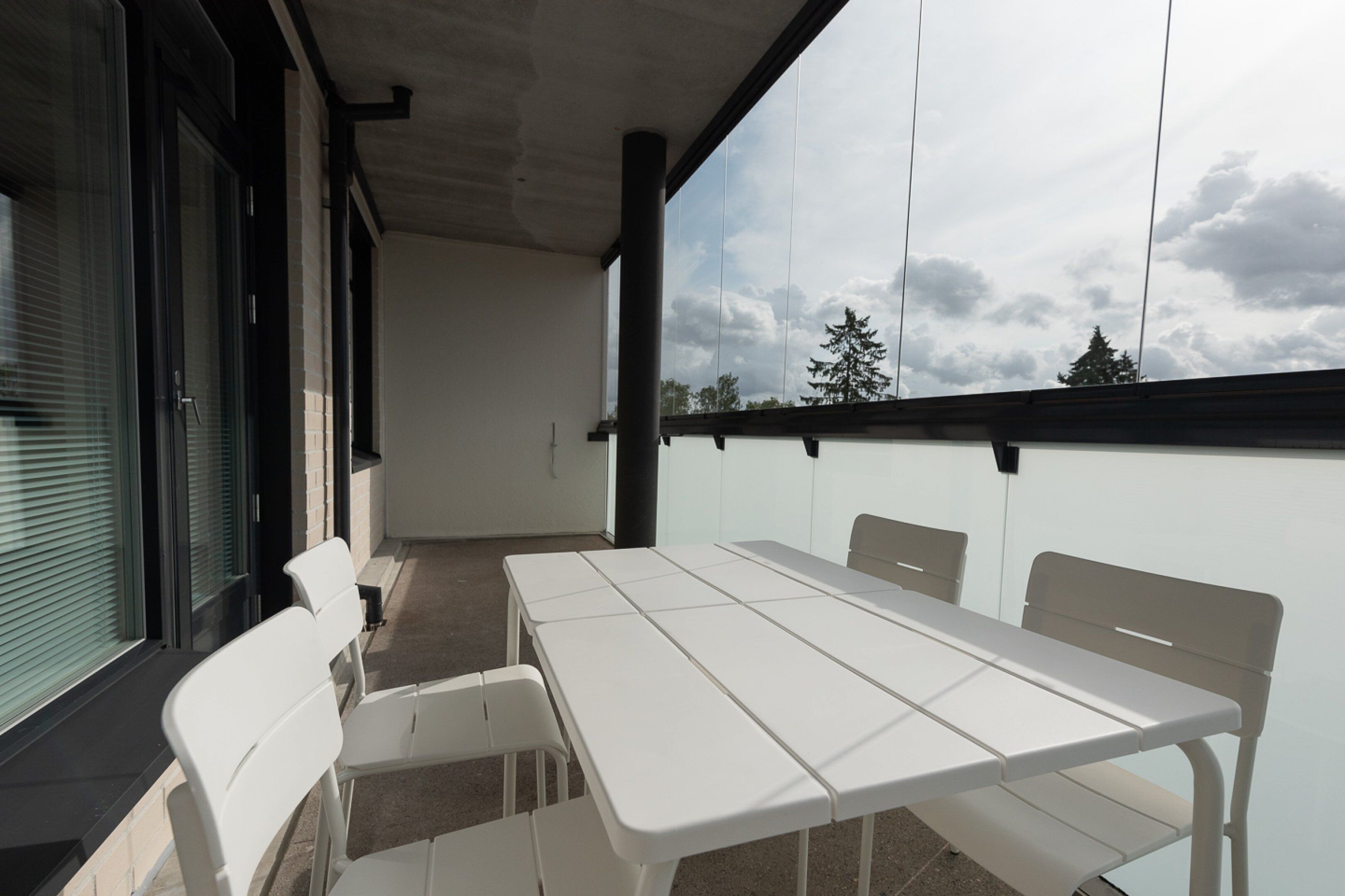 A balcony with a white table and chairs, a glass door, and a view of trees and sky.