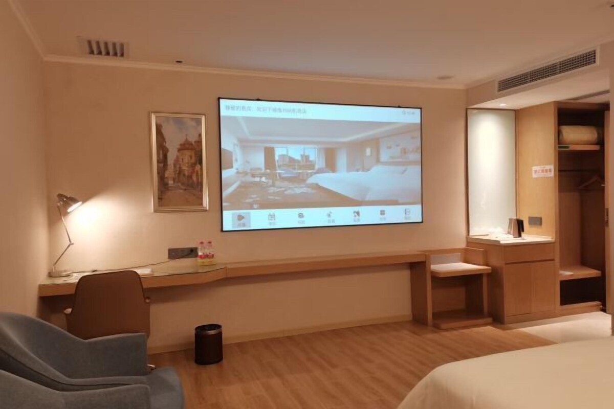 A hotel room with a bed, a desk, a chair, a projector screen displaying a room layout, and a wardrobe.