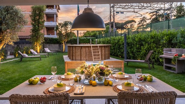 An outdoor dining area with a table set for a meal, surrounded by greenery and a view of a building in the background. - Egidio City Farmhouse (Lucca)