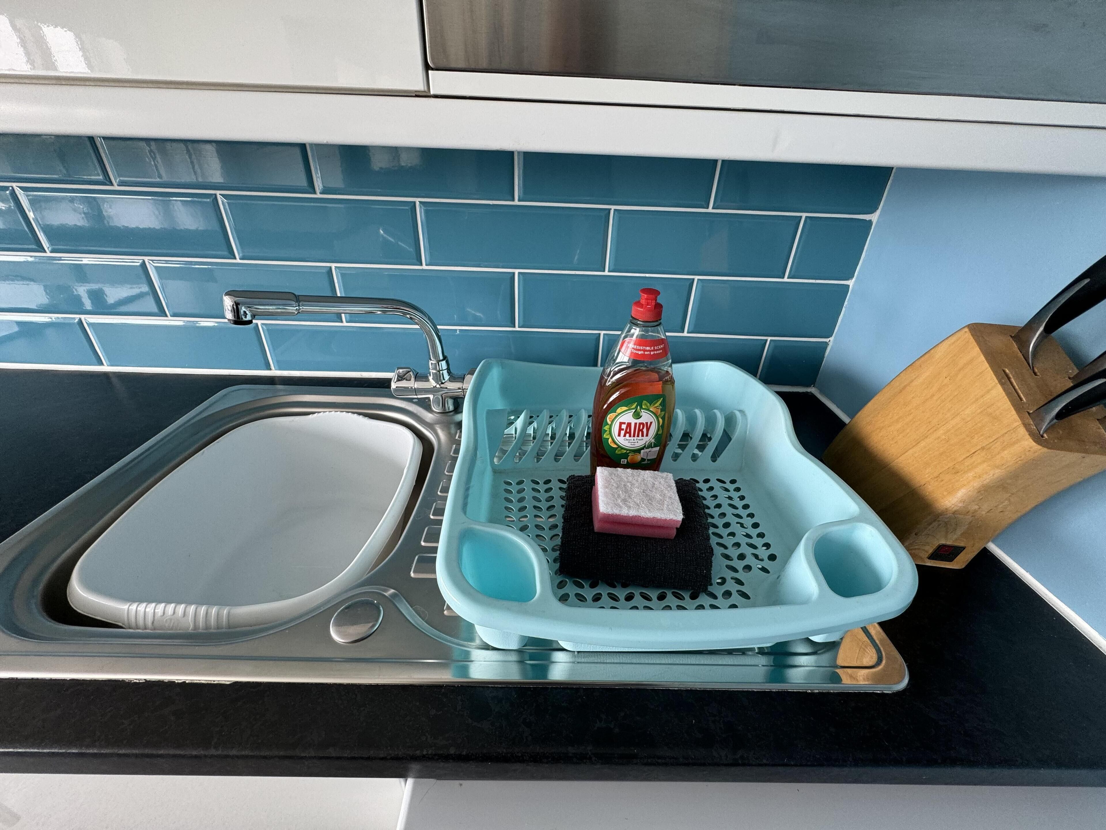 A kitchen with a sink, a dish drying rack, a bottle of Fairy dish soap, and a wooden cutting board.