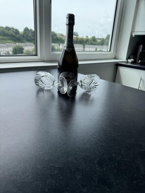 A bottle of Martini wine on a dark countertop with two crystal glasses, a window with a view of greenery and buildings, and a kitchen area in the background.
