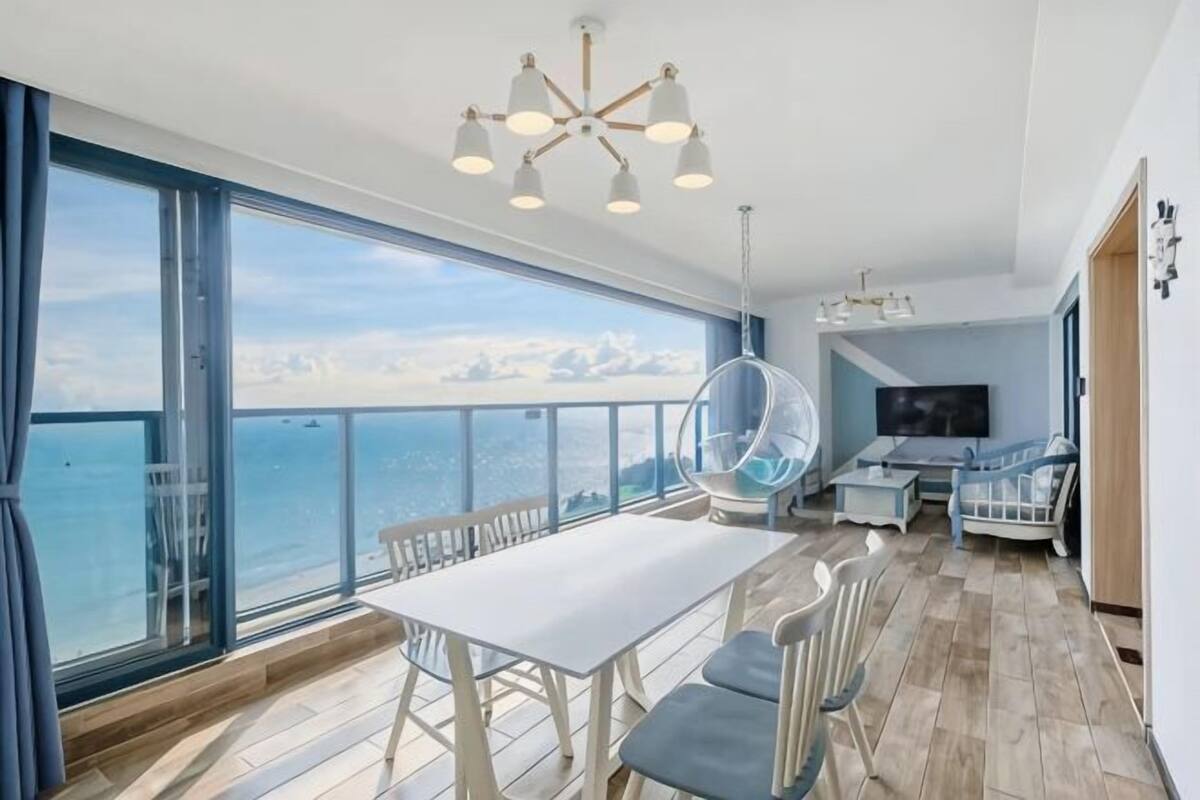 Una habitación moderna con un ventanal grande, una mesa de comedor, sillas, una butaca colgante, un televisor y suelo de madera.