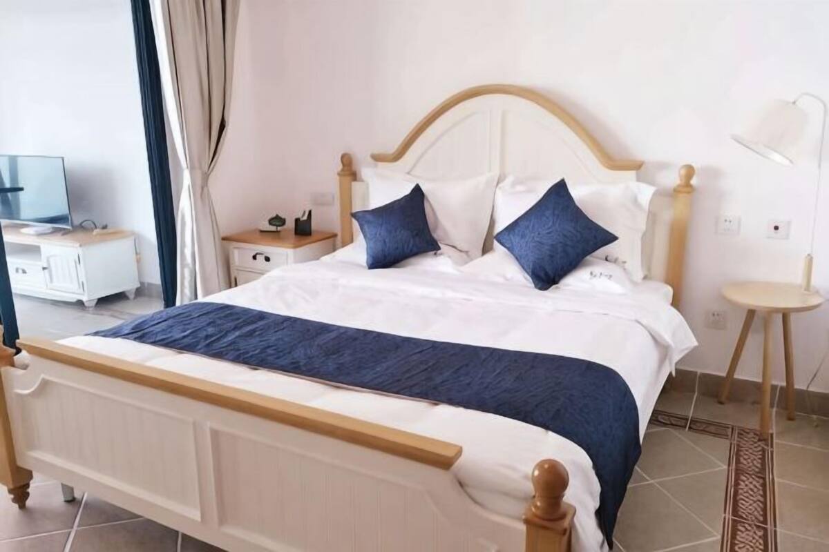 Una cama con estructura de madera, ropa de cama blanca y dos almohadas azules. Hay una mesita de noche con una lámpara y una pequeña mesa con otra lámpara.