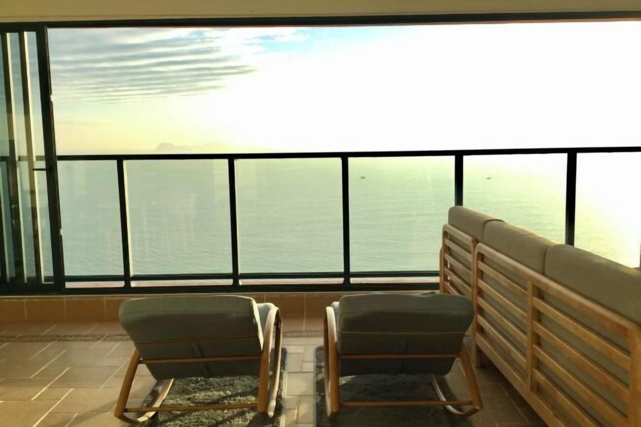 Quarto com uma grande janela com vista para um corpo d'água, duas cadeiras e um banco de madeira.