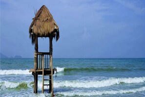 Une structure en bois, surmontée d’un toit de chaume, se dresse sur une plage, offrant une vue sur la mer.