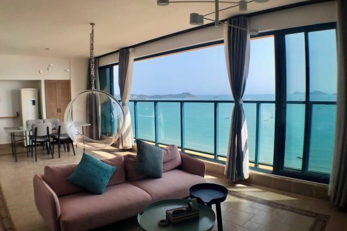 Ein modernes Wohnzimmer mit großem Fenster mit Blick auf das Meer, einer Couch mit Kissen, einem Hängesessel und einem kleinen Tisch.