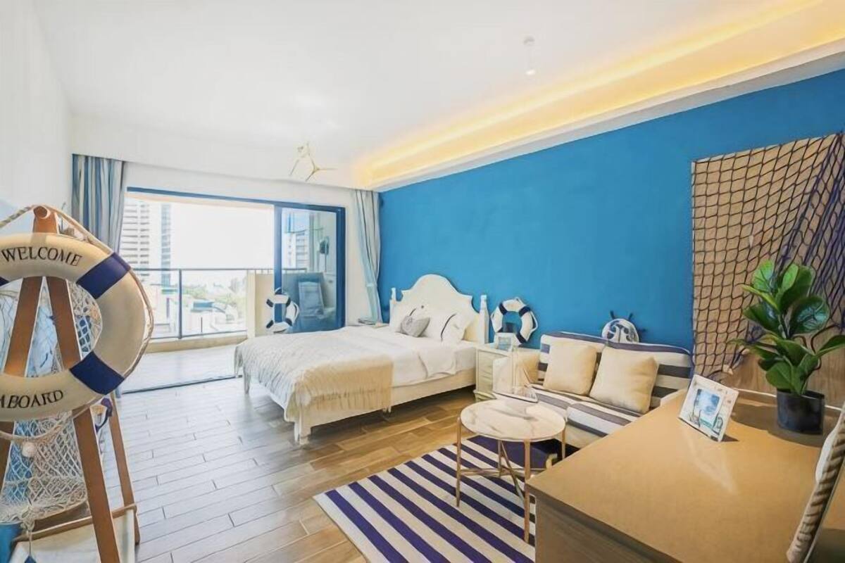 Habitación de hotel con cama, mesita de noche, un pequeño sofá, escritorio, silla, planta en maceta y balcón con vistas.