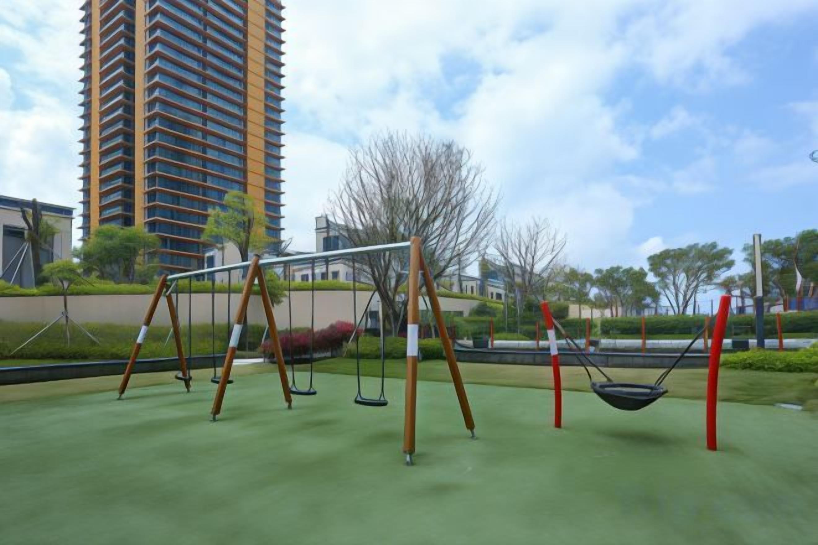 A playground with swings and a hammock, surrounded by greenery and a tall building in the background.