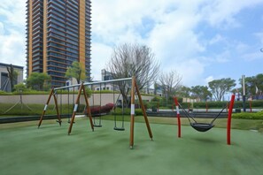A playground with swings and a hammock, surrounded by greenery and a tall building in the background.