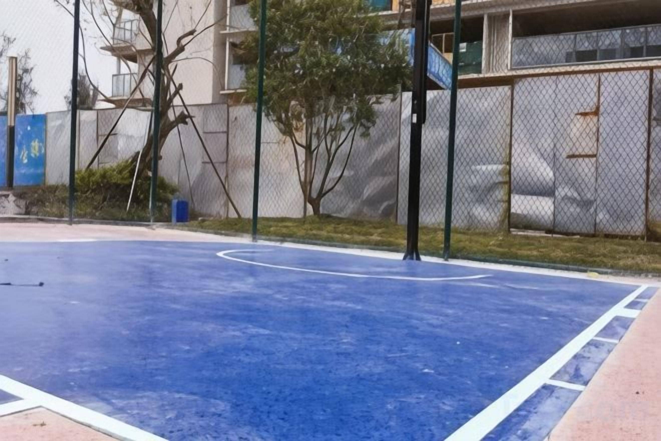 An outdoor basketball court with a blue surface, surrounded by a chain-link fence and trees.