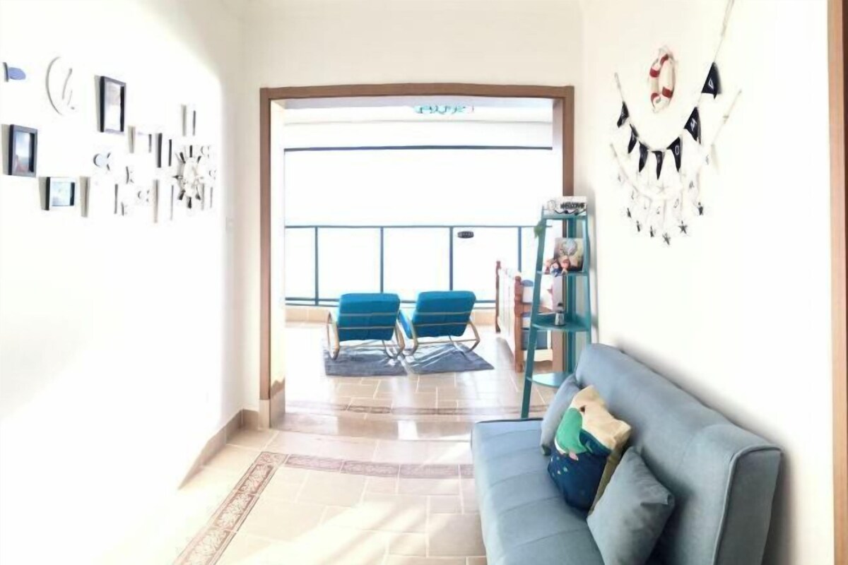Ein Wohnzimmer mit einem blauen Sofa, einem kleinen Tisch und einer Wanddekoration. Vor der Tür befinden sich zwei blaue Stühle.