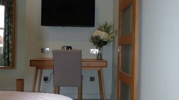 A bedroom with a bed, a desk with a chair, a television mounted on the wall, and a wooden door.