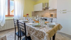 A dining area with a table set for four, a window with curtains, and a kitchen in the background. - Casa Clara Uri in the Nuraghe of Santa Cadrina a short distance from the beaches (Uri)