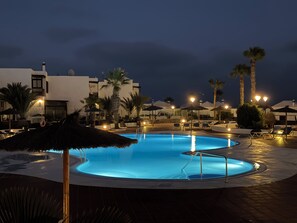 A swimming pool with a curved edge, surrounded by lounge chairs and umbrellas, with palm trees and a building in the background. - Casa Lyrata (Costa Teguise)