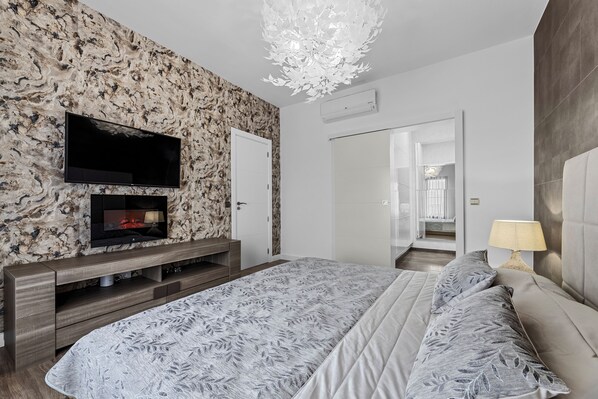 Un dormitorio moderno con una cama grande, un televisor de pantalla plana montado en una pared con textura, una mesita de consola de madera y una araña.