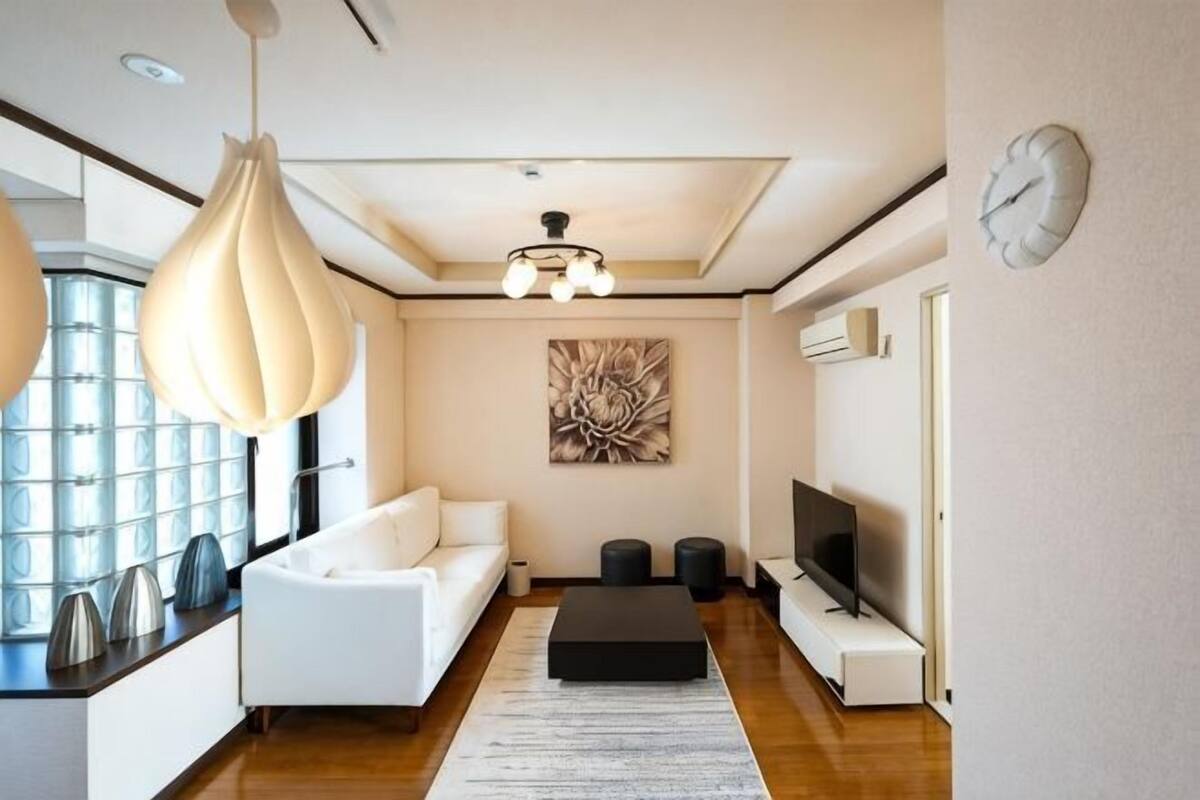 A modern living room with a white sofa, a black ottoman, a large abstract painting, a TV, and a clock on the wall.