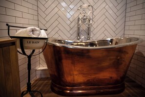 A copper bathtub with a towel on a rack labeled "LE BAIN" in a bathroom with white herringbone-patterned tiles.