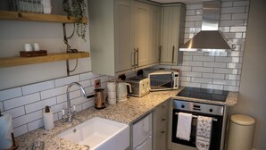 A compact kitchen with a sink, stove, and microwave. There are wooden shelves above the sink and a hanging plant. - High Spec 1-bed Cottage in Staindrop, Darlington (Darlington)
