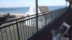 Ein Balkon mit Blick auf das Meer, einen Strand und einen Steg. Zwei weiße Stühle und ein Tisch mit einem Buch und einer Tasse stehen bereit.