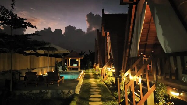 Vista nocturna de un complejo turístico con piscina, terraza de madera y un pabellón.