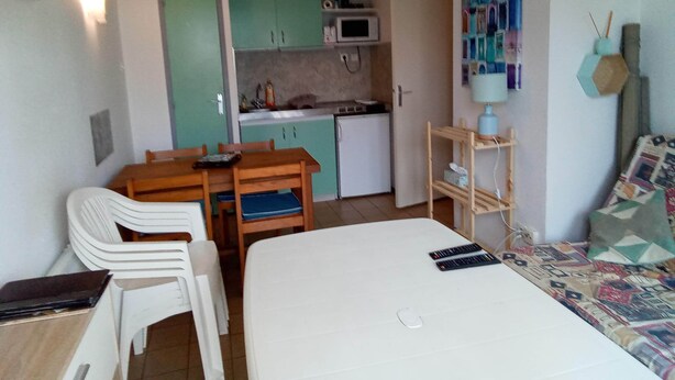 A compact room with a bed, a small dining table with chairs, a kitchenette, and a wall-mounted shelf with a lamp. - Au Port,agreable Appartement de Plain Pied Avec Terrasse (Hourtin)