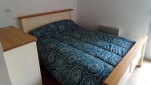 A single bed with a patterned bedspread in a room with plain walls and a radiator. - Au Port,agreable Appartement de Plain Pied Avec Terrasse (Hourtin)