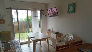A room with a dining table, chairs, a TV mounted on the wall, a clock, and a sliding glass door leading to a balcony. - Au Port,agreable Appartement de Plain Pied Avec Terrasse (Hourtin)
