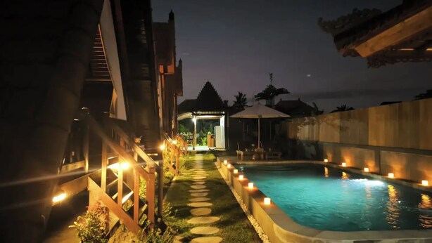 A nighttime view of a swimming pool area with a pathway lined with candles, a wooden structure, and a building in the background. - Lumbung Sari Ubud 2

 (Bali)