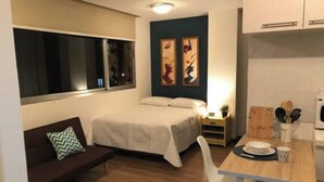Une pièce de taille modeste comprenant un lit, une petite table avec une assiette et des couverts, un canapé avec un coussin et une fenêtre.