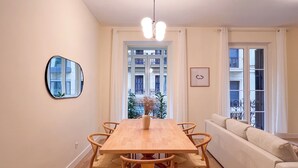 A dining area with a wooden table and chairs, a sofa, a mirror, and a view of buildings through the window.