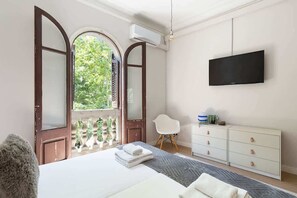 Un dormitorio con una cama grande, un televisor montado en la pared, un tocador blanco y una ventana con vistas a zonas verdes.