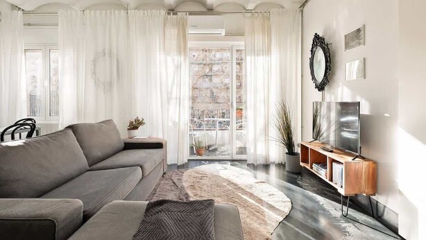 Una sala de estar moderna con un sofá gris, un mueble de televisión de madera y un ventanal con cortinas blancas.