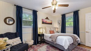 A bedroom with a bed, a nightstand, a chair, a ceiling fan, and a large window with curtains. - Chic Southern Retreat (Charleston)