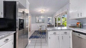 A modern kitchen with white cabinetry, a stainless steel refrigerator, a dishwasher, and a dining area with a blue patterned rug. - Chic Southern Retreat (Charleston)