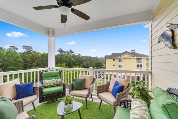 A covered outdoor seating area with wicker furniture, a ceiling fan, and a view of a yellow building in the background.