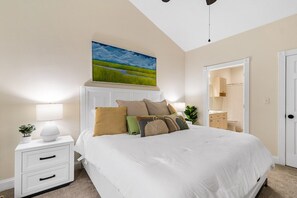 Quarto com uma cama grande, mesas de cabeceira brancas, um quadro acima da cama e uma porta que dá para o banheiro.