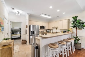 A modern kitchen with a stainless steel refrigerator, a microwave, and a built-in oven. There are white bar stools and a small island with stools.