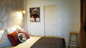 A bedroom with a bed, pillows, a wall-mounted picture, a door, and a small stool. - L'appart D'angélique (Imling)