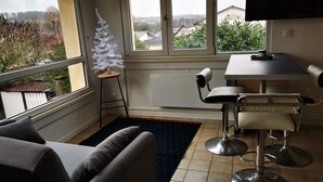 A modern living room with a sofa, a small table, and two bar stools. A decorative white tree is placed on a stool near the window. - L'appart D'angélique (Imling)