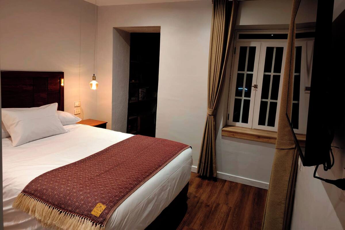 Ein Hotelzimmer mit einem großen Bett, einem Nachttisch mit Lampe, einem Fenster mit Vorhängen und einem Holzboden.